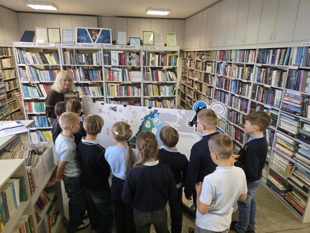 Nuotraukoje matyti grupė vaikų, stovinčių prieš didelį žemėlapį. Žemėlapis yra interaktyvus, jame matomi įvairūs piešiniai, susiję su šalimis ar veikėjais. Vaikai klausosi suaugusios moters, kuri rodo žemėlapį, ir aktyviai dalyvauja veikloje. Vaikai dėvi mokyklinius drabužius – mėlynus marškinėlius ir tamsias kelnes, o aplink juos yra gausios bibliotekos lentynos su knygomis. Atmosfera yra edukacinė ir žaisminga.