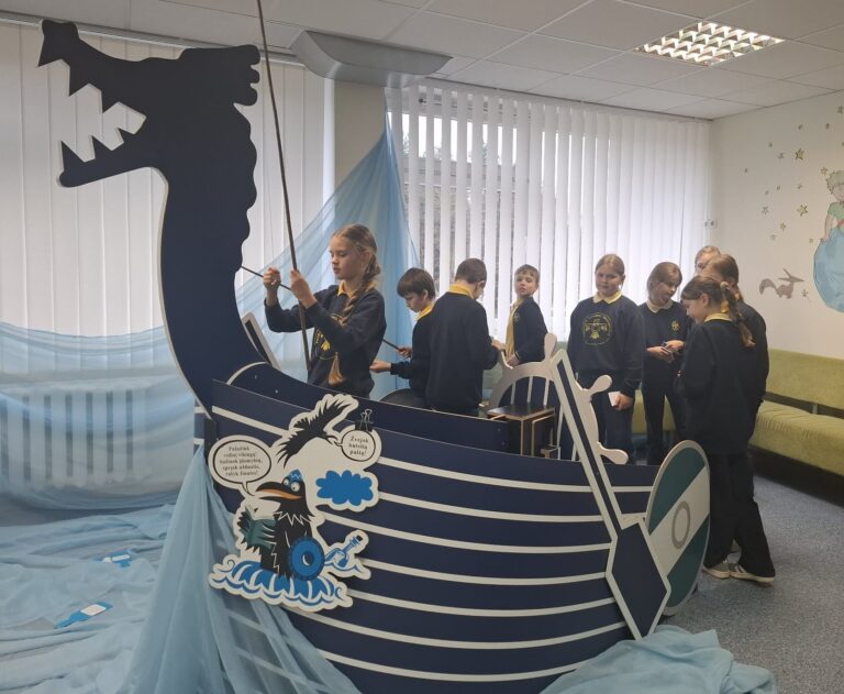 Klasėje keli pradinukai, vilkintys mokyklines uniformas, stovi ir žaidžia šalia didelės dekoratyvinės vikingų laivo instaliacijos su drakono galva; aplinka papuošta mėlynais audiniais, primenančiais jūrą.