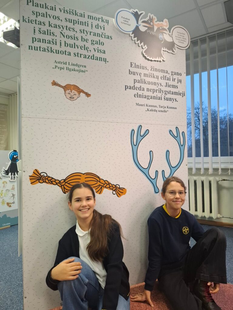 Nuotraukoje matomos dvi mergaitės, sėdinčios prie stendo, kuriame pateikiamos citatos iš žymių knygų. Stende iliustruoti Pepės Ilgakojinės (Astrid Lindgren kūrinys) ir elnio tema (Mauri Kunnas ir Tarja Kunnas kūrinys) personažai. Viena mergaitė sėdi ant grindų ir šypsosi, vilkėdama tamsų džemperį su geltonu Vyturio progimnazijos logotipu, o kita sėdi šalia ir taip pat šypsosi, dėvėdama juodą džemperį su baltais marškiniais.