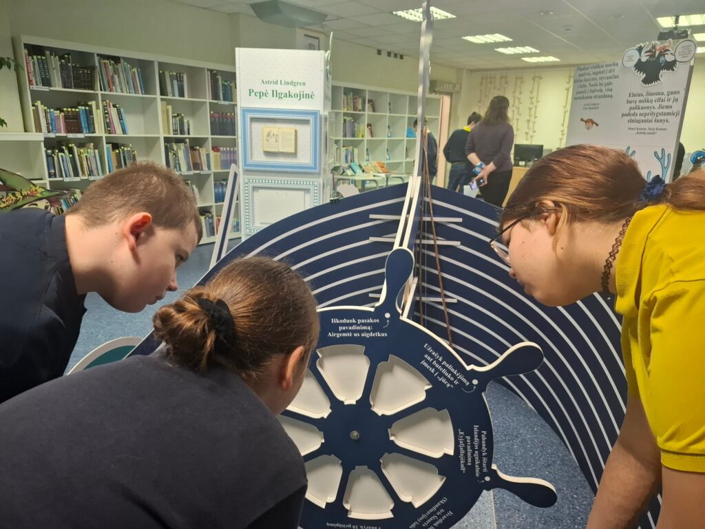 Bibliotekoje trys vaikai pasilenkę apžiūri interaktyvų ekspozicijos ratą, susijusį su Astridos Lindgren knyga „Pepė Ilgakojinė“; fone matyti knygų lentynos ir kiti lankytojai.