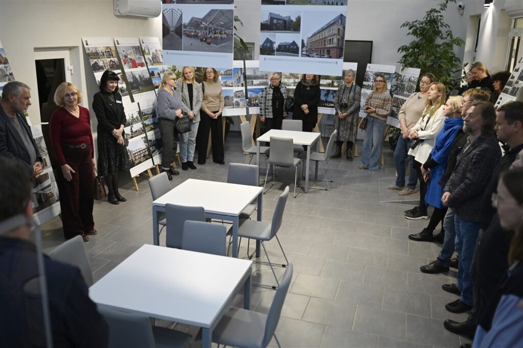 Patalpoje išdėstyti dideli nuotraukų stendai, kuriuose matomi architektūriniai projektai ar pastatai. Grupė žmonių, tiek vyrų, tiek moterų, stebi šias parodas, kai kurie diskutuoja tarpusavyje. Aplinka moderni ir tvarkinga, su baltomis sienomis, pilkšvais grindimis ir paprastu baldų dizainu. 