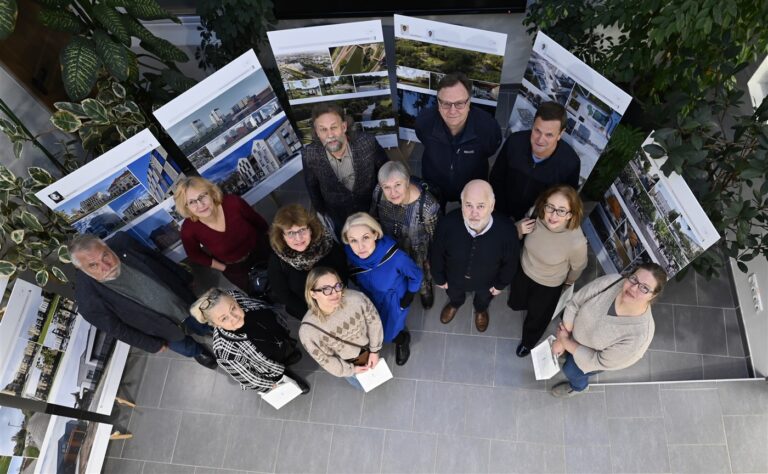 Iš viršaus fotografuota grupė žmonių stovi ratu tarp didelių architektūrinių vizualizacijų stendų parodoje; jie žvelgia į kamerą šviesioje, augalais apželdintoje erdvėje.