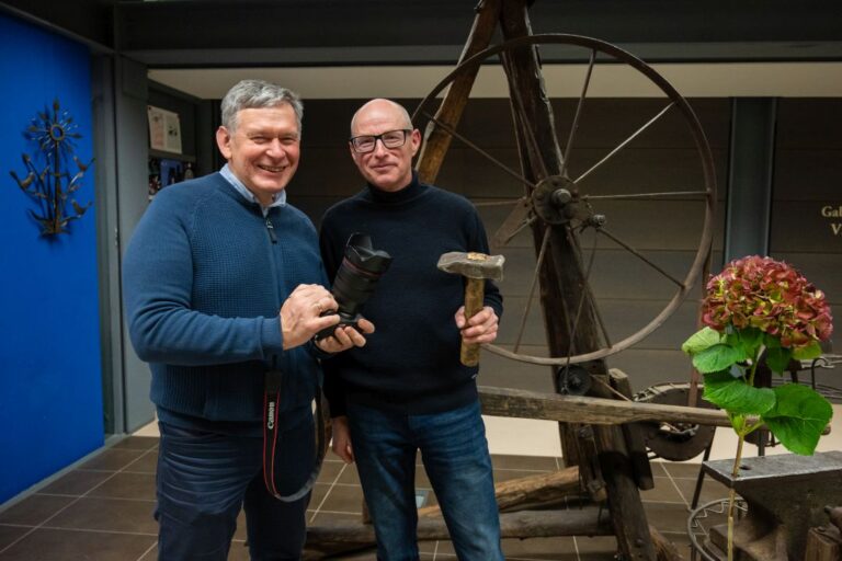 Stovi ir šypsosi du vyrai, vienas su fotoaparatu rankoje, kitas su kalvio įrankiu kūju.