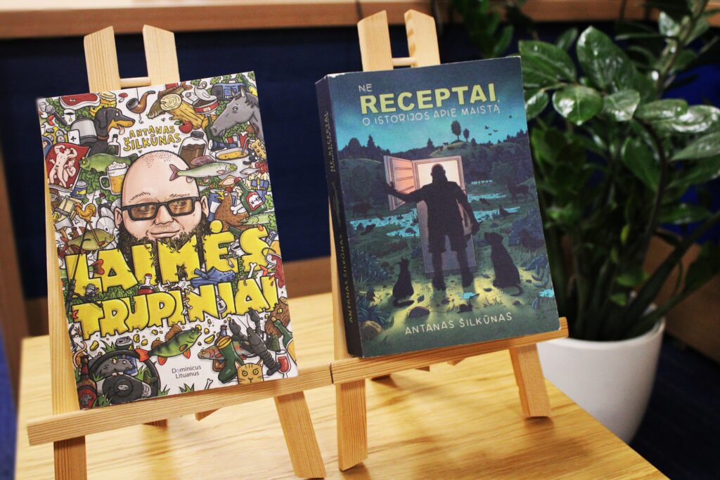 Nuotraukoje matomos dvi knygos, padėtos ant medinio stovo. Kairėje pusėje – knyga „Laimės trupiniai“ su spalvingu piešiniu viršelyje, kur vaizduojamas autorius Antanas Šilkūnas ir įvairūs daiktai. Dešinėje pusėje – knyga „Ne receptai, o istorijos apie maistą“, kurios viršelyje pavaizduotas žmogus, atidarantis duris į naktinę gamtą. Fone matoma žalia augalų dekoracija.