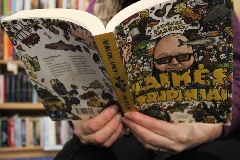 Nuotraukoje matoma knyga su spalvingu piešiniu viršelyje. Knygos pavadinimas – „Laimės trupiniai“, o autorius – Antanas Šilkūnas. Knyga atvira, ją laiko žmogus, kuris skaito. Fone matosi knygų lentynos. Viršelis iliustruotas įvairiais komiksais ir daiktais, įskaitant žuvį, alaus bokalus ir kitus smulkius elementus.