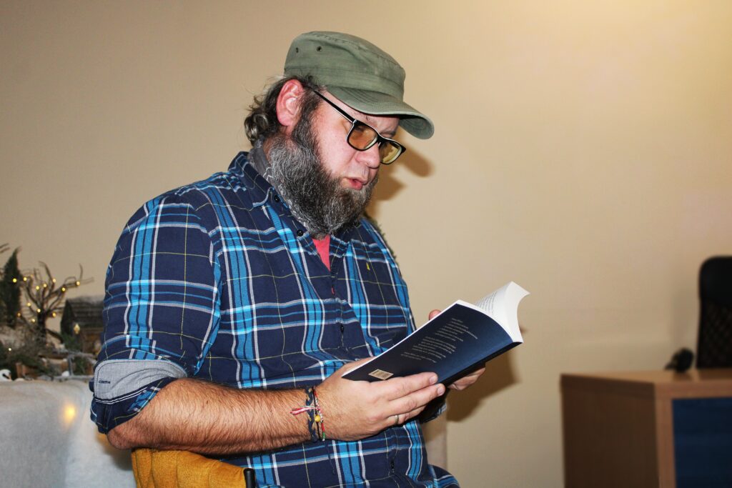 Nuotrauka rodo vyrą, skaitantį knygą. Jis dėvi mėlyną ir žalią languotą marškinį, o ant galvos turi žalią kepurę. Vyras turi pilką barzdą ir akinius. Jo rankoje matyti keli apyrankės, o ant stalo šalia yra kėdė. Fone matomi šviesos elementai, galintys būti kalėdine dekoracija.