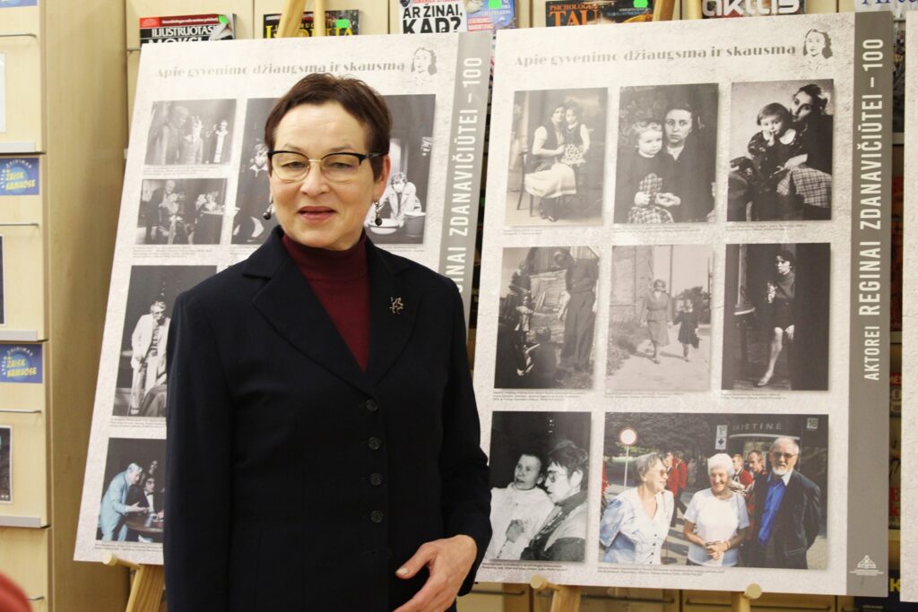 Moteris su akiniais ir tamsiu švarku stovi patalpoje prie dviejų stendų su senomis nespalvotomis ir spalvotomis nuotraukomis, skirtomis aktorės Reginos Zdanavičiūtės 100-mečiui.
