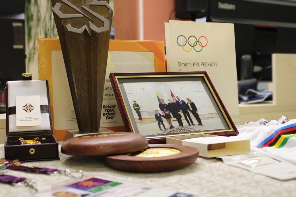 Nuotraukoje matomi įvairūs apdovanojimai, medaliai ir dokumentai, priklausantys Simona Krupėckaitei. Pirmoje plano dalyje matome medalius ir apdovanojimus, įskaitant apdovanojimų dėžutę su ženkliukais. Taip pat yra diplomai ir sertifikatai, įskaitant Olimpinio komiteto dokumentą, kuriame paminėtas Simona Krupėckaitė. Nuotrauka taip pat vaizduoja šeimą ir jos susitikimą su valdžios atstovais, matant jų nuotrauką su prezidentu ir kitais asmenimis.