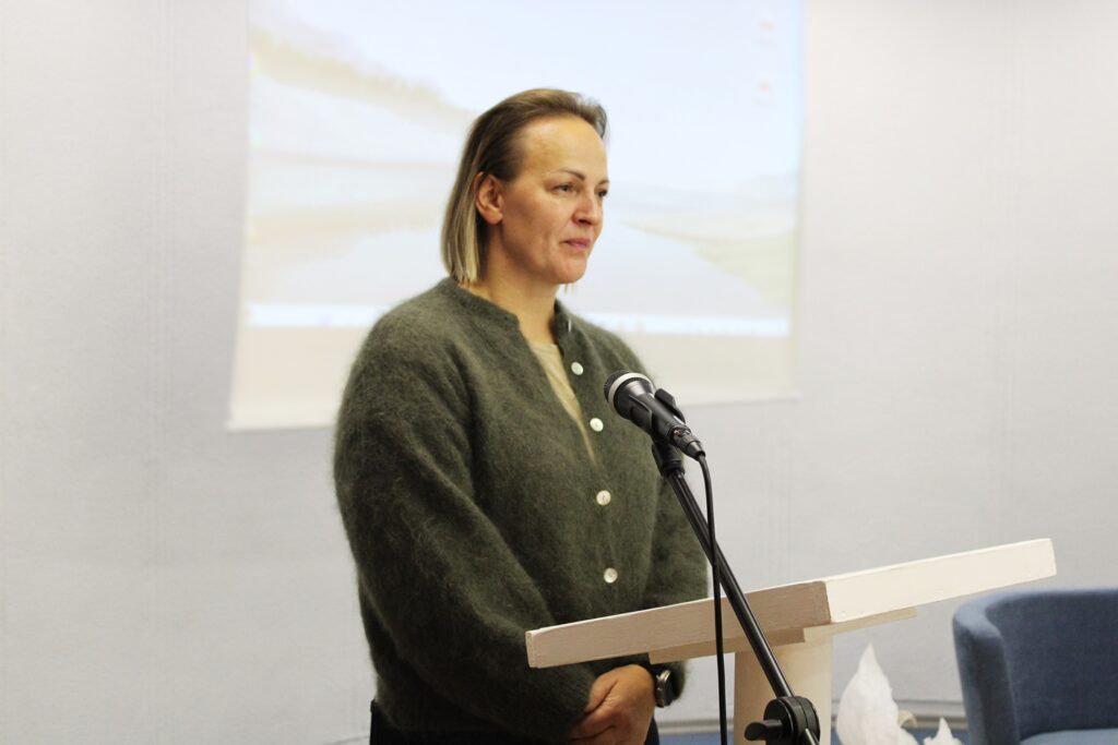 Simona Krupeckaitė stovi prie tribūnos ir kalba į mikrofoną, už jos matomas projekcinis ekranas.