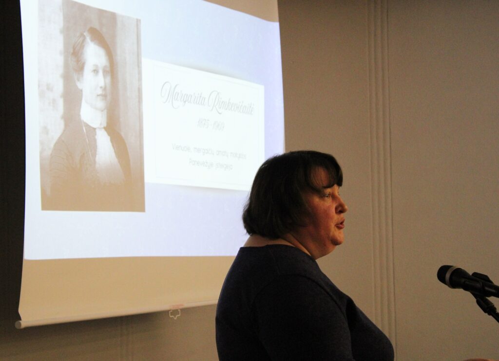 Nuotraukoje matoma moteris, kalbanti prie mikrofono, o už jos ekrane rodomas senas portretas. Ekrane matome informaciją apie Margaritą Rimkevičiūtę (1875–1969), paminint jos gyvenimo metus ir vaidmenį kaip vieną iš pirmųjų moterų, dirbusių Panevėžio gydytojų tarpe.
