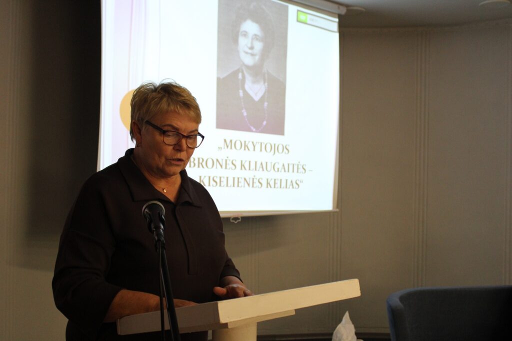 Nuotraukoje matoma moteris, kalbanti prie podiumo, o už jos ekrane rodomas senas portretas. Ekrane taip pat matoma frazė „Mokytojos Bronės Kliugaitės – Kiselines kelias“.