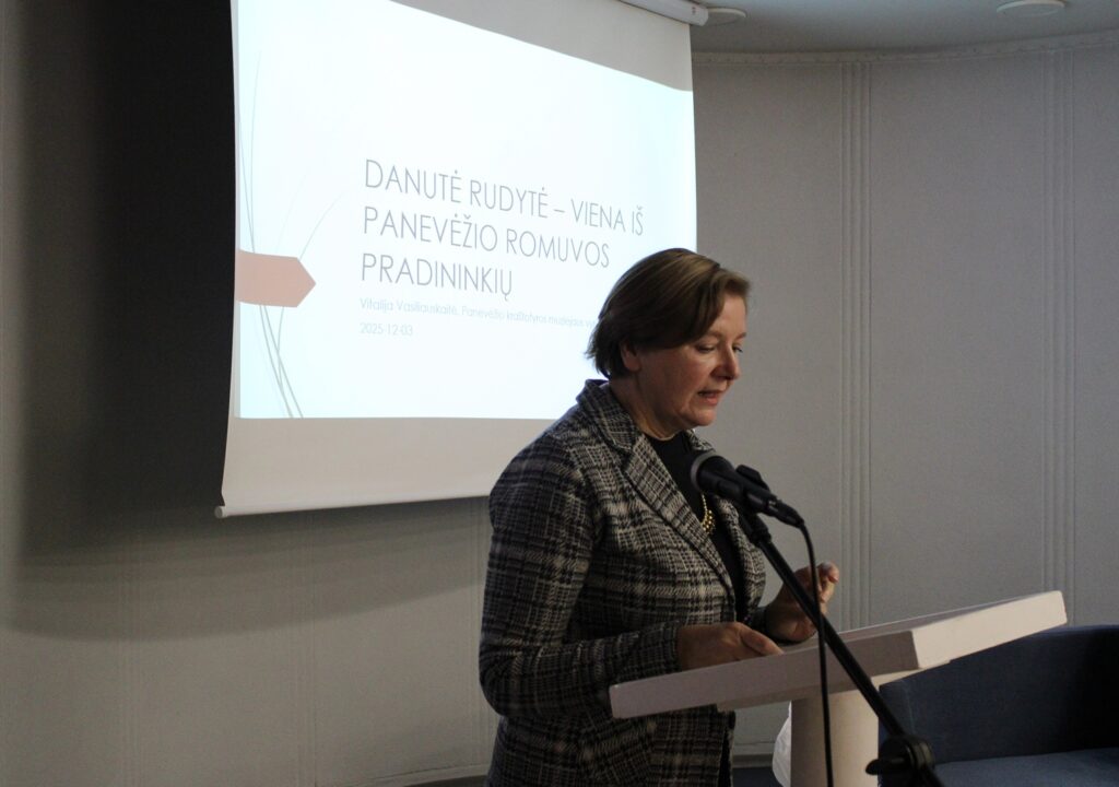 Nuotraukoje matoma moteris, kalbanti prie podiumo, o už jos ekrane rodomas pranešimas, kuriame paminima „Danutė Rūdytė – viena iš Panevėžio Romuvos pradininkių". Pranešimą skaito Vitalija Vasiliauskaitė, pristatanti šią asmenybę Panevėžio krašto istorijoje, su renginio data – 2025 m. gruodžio 3 d.