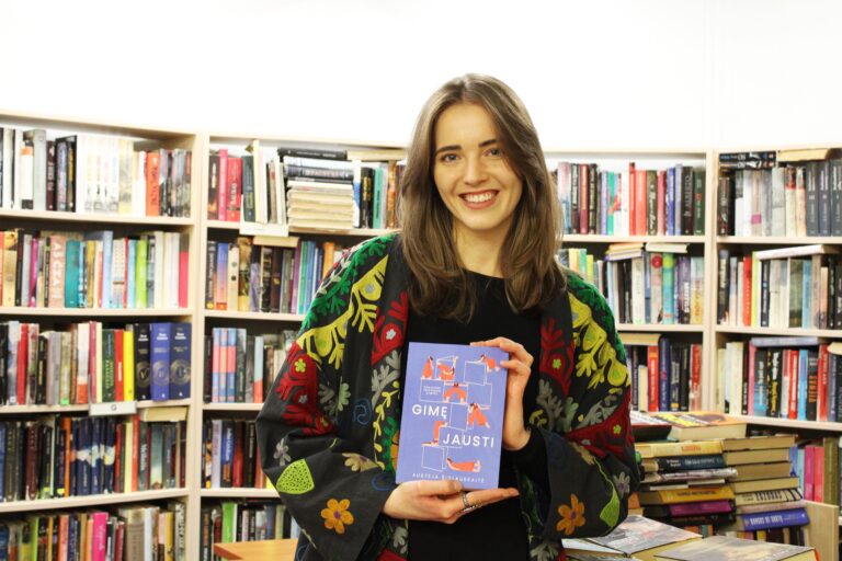 Šypsodamasi moteris knygyne laiko rankose knygą violetiniu viršeliu, stovėdama priešais lentynas, pilnas įvairių knygų.