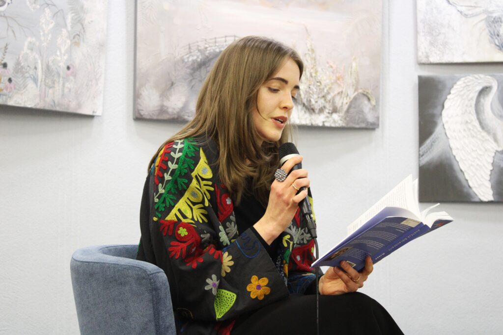 Ant kėdės sėdinti moteris spalvingu raštuotu apsiaustu garsiai skaito iš atverstos knygos, laikydama mikrofoną. Fone matyti švelnių tonų paveikslai.