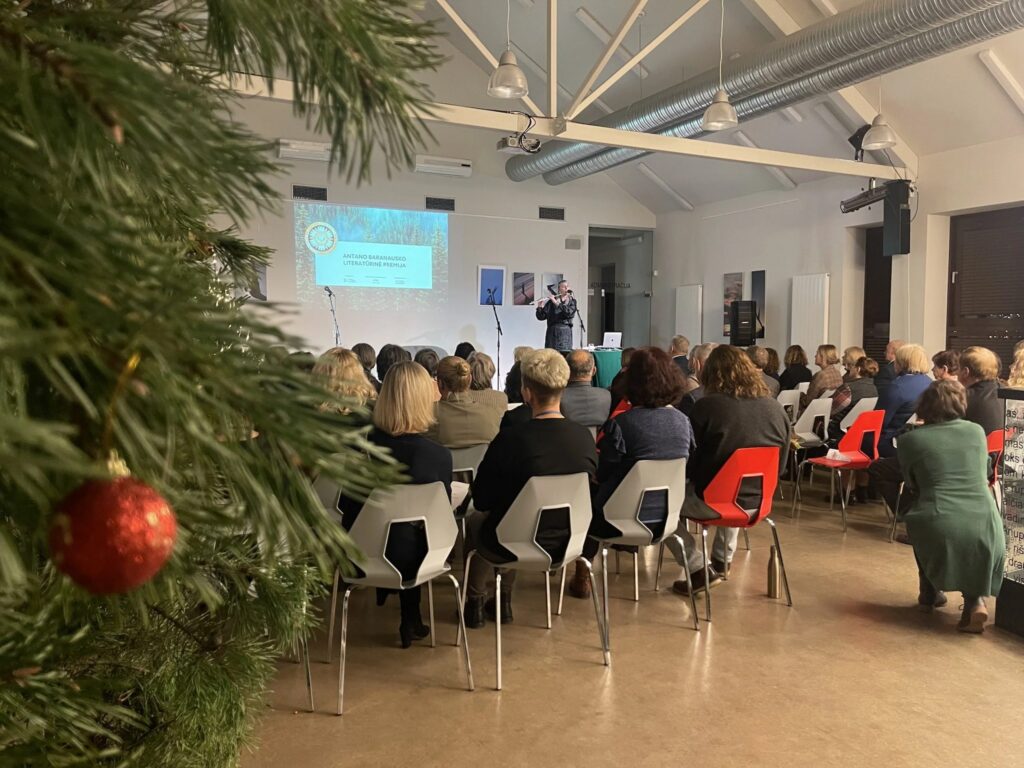Nuotraukoje matoma apdovanojimo ceremonija. Pirmame plane – Kalėdų eglutė su raudona dekoracija, o fone – daug žiūrovų, sėdinčių salėje ir stebinčių renginį. Prie mikrofono kalba žmogus, o ekrane matoma informacija apie Antano Baranausko literatūrinę premiją. Salė šviesi, moderni, su dideliais langais, o žiūrovai sėdi ant baltų ir raudonų kėdžių.