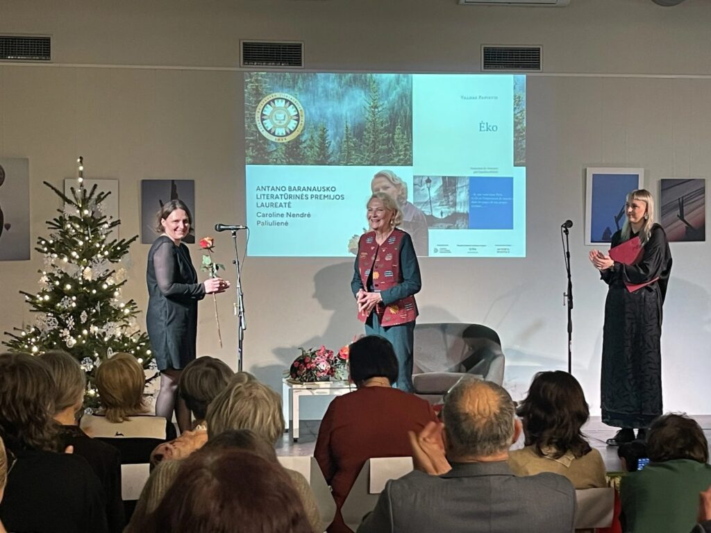 Nuotraukoje matomas apdovanojimo ceremonijos momentas. Centre stovi Caroline Nendrė Paliulienė, laikanti raudoną rožę ir šypsosi, o šalia jos – dvi moterys, viena iš jų ploja, o kita sveikina su apdovanojimu. Fone matoma kalėdų eglutė su dekoracijomis, taip pat ekrane rodomas apdovanojimas ir laureatės pavardė. Žiūrovai sėdi ir stebi ceremoniją.