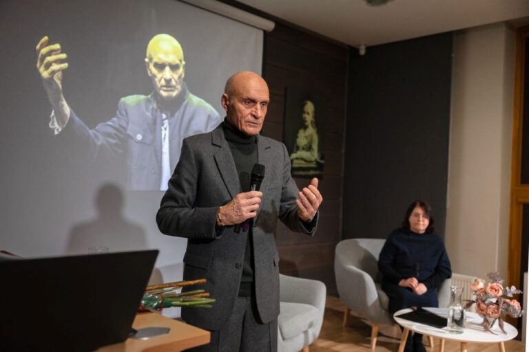 Vyras išraiškingai laiko rankas su mikrofonu vienoje rankoje salės fone