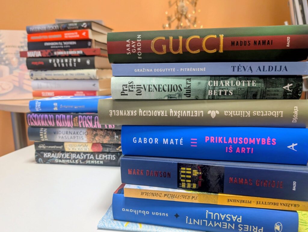 Ant stalo sukrauta knygų krūva su įvairių autorių leidiniais; matyti grožinės literatūros ir negrožinių knygų viršeliai, fone – šventinė aplinka.