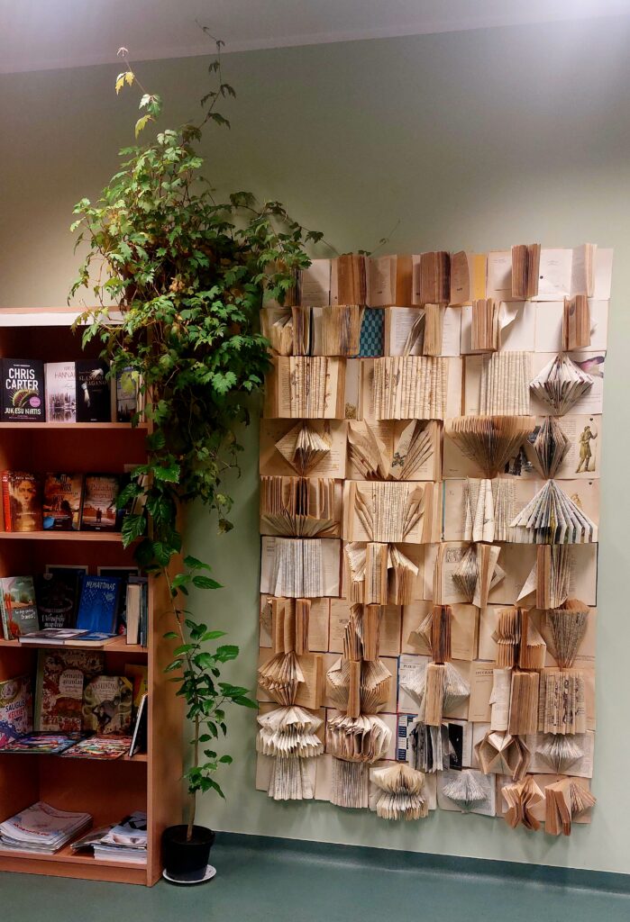 Nuotrauka rodo erdvę su įdomiu sienos dekoru, kuris sukurtas iš sulankstytų ir išdėliotų knygų puslapių. Knygų puslapiai suformuoti į įvairius meno kūrinius, pvz., trikampius, lankus ir kitus geometrinius bei organinius raštus. Sienos dešinėje pusėje matome augalą, auginamą vazone, kuris prideda natūralios žalumos. Kairėje pusėje yra lentyna su įvairiomis knygomis ir žurnalais. Ši erdvė atrodo jauki ir kūrybinga, puikiai dera su knygų tematika.