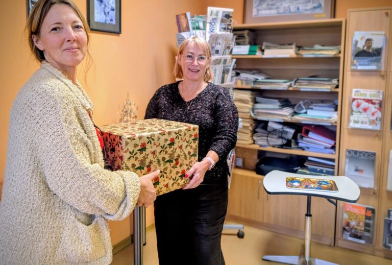 Dvi moterys bibliotekoje perduoda viena kitai dovanų supakuotą dėžę; už jų matyti lentynos su knygomis ir žurnalais, šalia stovi mažas staliukas su saldumynais.