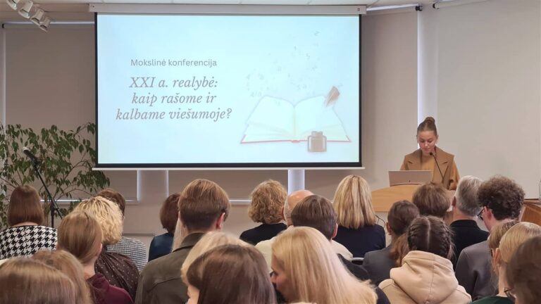 Mokslinės konferencijos salėje sėdi gausi auditorija, žiūrinti į pranešėją, stovinčią prie tribūnos dešinėje. Ant didelio projekcinio ekrano rodomas konferencijos pavadinimas „XXI a. realybė: kaip rašome ir kalbame viešumoje?“ bei atverstos knygos iliustracija.