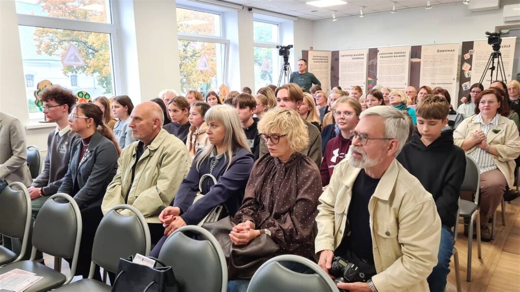 Salėje sėdi gausi įvairaus amžiaus auditorija, susitelkusi ir klausanti renginio. Priekyje matomi kelių eilių žiūrovai, o fone – informaciniai stendai ir filmavimo įranga.