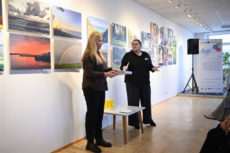 Nuotraukoje matyti parodos atidarymas, kuriame dvi moterys kalba ir dalijasi informacija su auditorija. Pirmoji moteris laiko dėžutę, o antroji kalba, tikėtina, pristatydama parodą. Už jų ant sienos pakabintos įvairios nuotraukos, vaizduojančios gamtą ir žmones. Kairėje matoma garso aparatūra, o dešinėje – stendas su informacija apie parodą. Renginys vyksta modernioje galerijoje, su šviesia aplinka ir medinėmis grindimis.