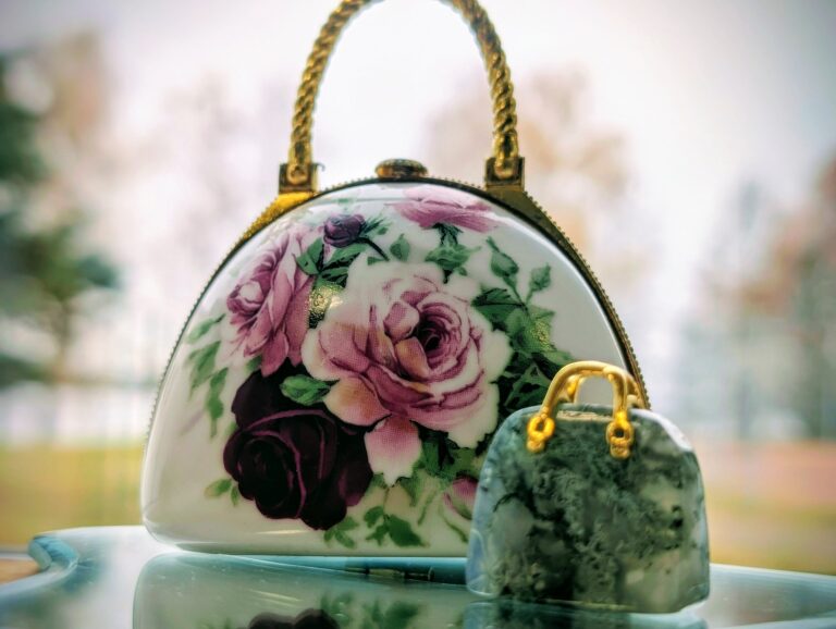 Nuotraukoje matome du miniatiūrinius rankinukus. Pirmasis rankinukas, esantis kairėje pusėje, yra baltas su rožinių ir tamsiai raudonų rožių gėlėmis, dekoruotas aukso spalvos rankena. Antrasis, mažesnis rankinukas, yra pilkšvas ir pagamintas iš akmens, su aukso spalvos rankena. Pagrindas, ant kurio padėti rankinukai, atrodo šviesus ir šiek tiek blizgus, o fonas neryškus su šviesiais medžiais.