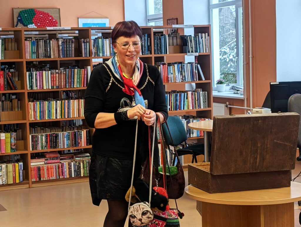 Nuotraukoje matome moterį, stovinčią bibliotekos aplinkoje, kurioje matosi knygų lentynos. Ji šypsosi ir laiko kelis miniatiūrinius rankinukus, pakabintus ant peties. Moteris dėvi juodą megztinį su spalvotu šalikėliu ir juodą sijoną. Pagrindiniame plane taip pat matosi medinis dėžutė, stovi ant stalo. Biblioteka turi šiltą ir jaukią atmosferą, o pro langą matosi sniegas.