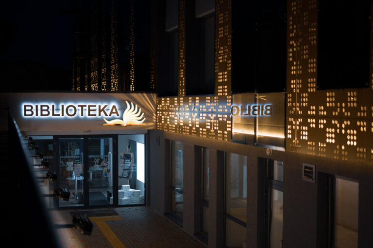 Vakare apšviestas šiuolaikinis bibliotekos įėjimas su šviečiančiu užrašu „Biblioteka“ ir stilizuotos atverstos knygos logotipu; šalia matyti perforuotas sienų apšvietimas bei stiklinės durys į vidų.