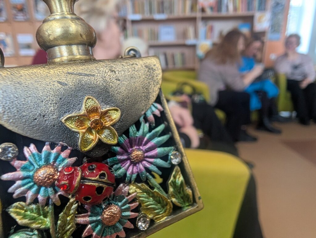 Nuotraukoje matome detalią miniatiūrinio rankinuko, puošto gėlėmis ir boružėle, nuotrauką. Rankinukas pagamintas iš metalo, o jo paviršius dekoruotas spalvotomis gėlėmis ir akmenukais, su ryškiai raudona boružėle centre. Fone matosi grupė žmonių, sėdinčių ant žalių sofų, o už jų yra bibliotekos lentynos su knygomis. Atmosfera atrodo šilta ir jauki, o žmonės bendrauja.