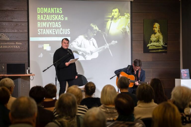 Priešais auditoriją stovi poetas ir skaito iš knygos, šalia su gitara sėdi bardas salės fone
