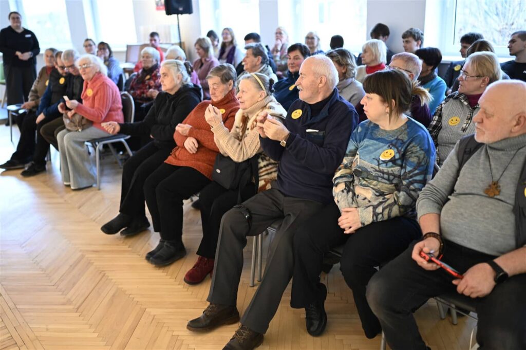 Nuotraukoje matyti renginio dalyviai, sėdintys salėje ir stebintys renginį. Žmonės yra įvairaus amžiaus, o daugelis jų dėvi geltonus lipdukus su šypsenėlėmis. Patalpa šviesi, su medinėmis grindimis ir daugybe langų. Pirmoje eilėje matome kelis vyresnio amžiaus žmones, kurie, atrodo, žiūri į priekį arba bendrauja tarpusavyje. Įvykio atmosfera šilta ir draugiška.