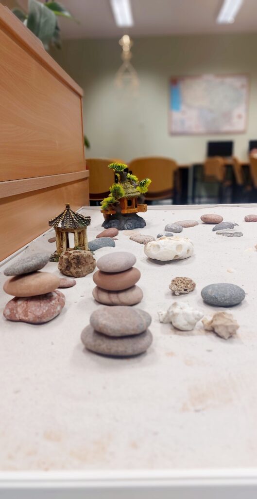 Nuotrauka vaizduoja zen sodą, kuriame yra įvairių akmenų, smėlio ir mažų dekoratyvinių elementų, tokių kaip miniatiūriniai paviljonai ir augalai. Akmenys sudėlioti į įvairias kompozicijas, sukuriant ramybės ir harmonijos įspūdį.