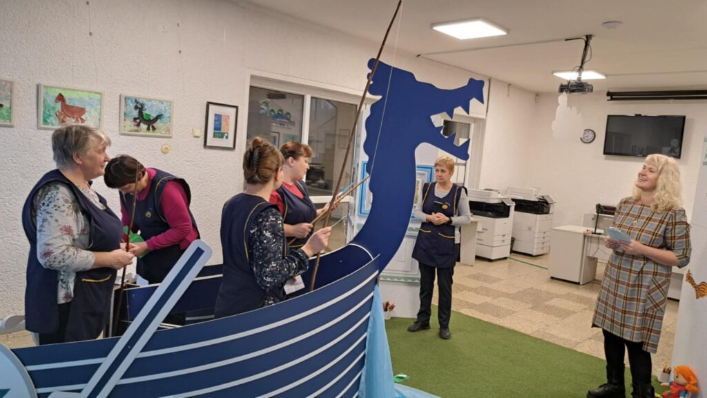 Nuotraukoje matome darbuotojas, kurios ruošiasi edukacinei veiklai. Jos stovi prie didelio laivo maketo, kurio priekyje matosi vikingų drakono galva. Kelios moterys užsiima paruošiamaisiais darbais: viena laiko žvejybinį kotą, o kita tvarko aplinką. Fone matome suaugusią moterį, kuri šypsosi ir kalba, galbūt pasakoja apie veiklą ar pasidalija instrukcijomis. Aplinka šviesi, su meno kūriniais ant sienos ir šiuolaikinėmis technologijomis. Bibliotekos grindys yra padengtos žalia kilimine danga, kuri sukuria jaukią ir edukacinę atmosferą.