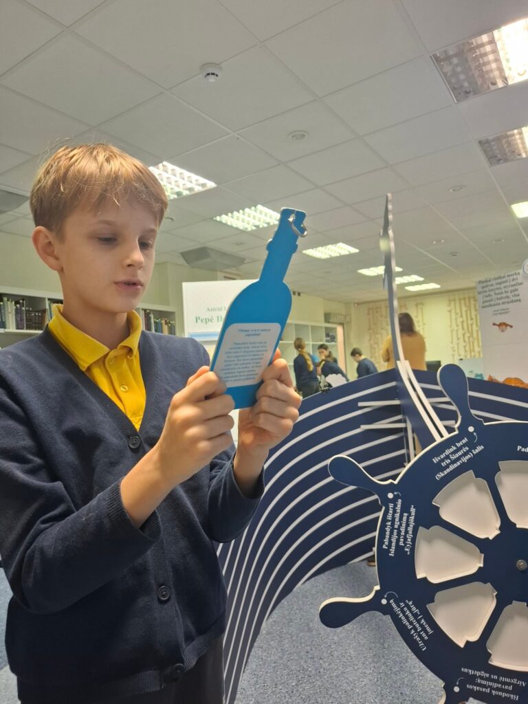 Nuotraukoje matomas berniukas, laikantis mėlyną kortelę, kurios forma primena butelio siluetą. Jis žiūri į kortelę, kurioje pateikiamas klausimas. Fone matomas stendas su įvairiais edukaciniais elementais ir žaismingais grafiniais elementais. Berniukas vilki tamsiai mėlyną džemperį su geltonais apykaklės kraštais, o aplinkui matosi kiti žmonės ir bibliotekos aplinka.