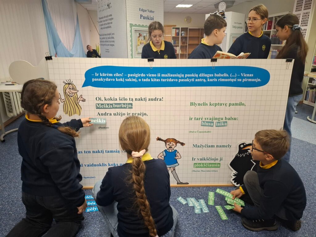 Bibliotekoje keli vaikai, vilkintys mokyklines uniformas, kartu dėlioja ir skaito eilėraščių ištraukas ant didelio interaktyvaus stendo su literatūriniais personažais; dalis vaikų klūpo, kiti stovi ir diskutuoja.