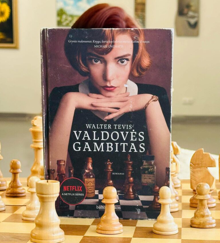 Knyga „Valdovės gambitas“ (Walter Tevis) pastatyta ant šachmatų lentos, priekyje matomos medinės šachmatų figūros. Ant viršelio – jaunos moters portretas, o virš knygos matoma tikro žmogaus šukuosena, sutampanti su viršelio nuotraukoje pavaizduotos moters šukuosena.