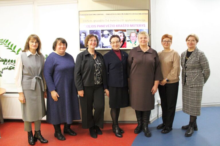 Septynios moterys stovi vienoje eilėje patalpoje prie sienos su ekranu. Jos šypsosi, vilki įvairių spalvų tvarkius drabužius. Fone matomas ekranas su konferencijos skelbimu apie iškilias Panevėžio krašto moteris.