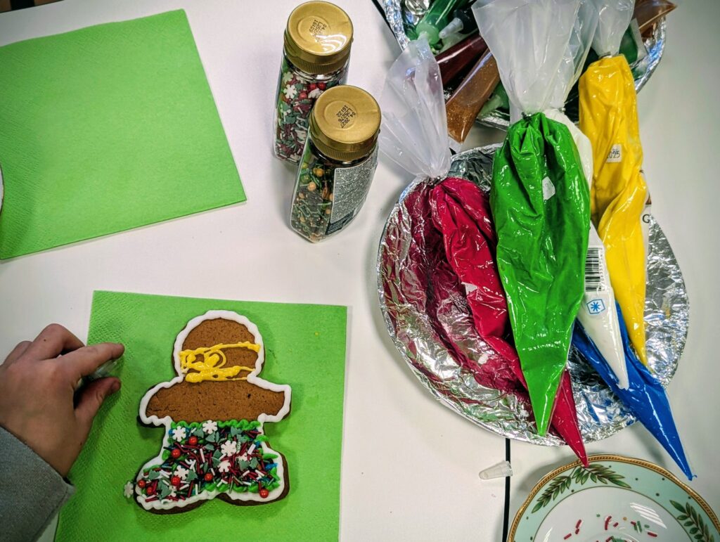 Nuotraukoje matomas puošiamas imbiero sausainis, atvaizduojantis žmogeliuką. Jis dekoruojamas balta ir geltona glajumi, taip pat papuoštas spalvotais konfeti ir saldainių žirneliais. Stalas padengtas žaliu servetėliu, o šalia matomi įvairių spalvų glajai maišeliuose ir indeliai su spalvotais dekorais.