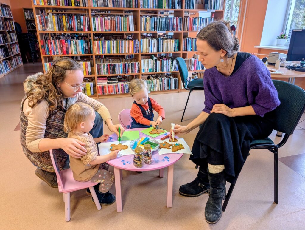Nuotraukoje matoma kūrybinė veikla bibliotekoje, kur mama ir du maži vaikai dekoruoja imbiero sausainius. Vaikai sėdi prie mažo rožinio staliuko, naudodami spalvotus dažus ir dekoravimo priemones. Mama, vilkinti purpurinę megztinę, sėdi šalia ir taip pat puošia sausainius. Fone matomos knygos ir bibliotekos interjeras.