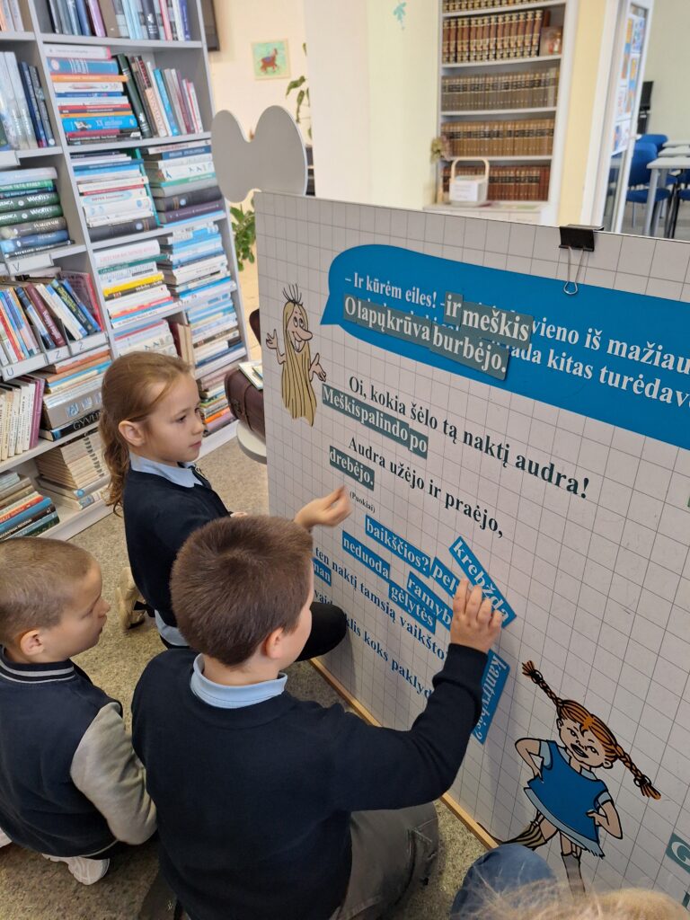 Nuotraukoje matyti grupė vaikų, kurie dalyvauja interaktyvioje veikloje, susijusioje su Tove Jansson kūriniais. Vaikai stovi šalia didelio plakato, ant kurio užrašytas tekstas iš knygos. Jie aktyviai prisideda prie užduoties, perkeldami tekstinius elementus į tinkamas vietas, galimai sudarydami ar keičiant sakinius. Plakate matomi piešiniai su troliais ir citatos, kurios susijusios su knygų veikėjais. Fone matomos bibliotekos lentynos su knygomis, kurios kuria šiltą ir edukacinę atmosferą. Vaikai dėvi mokyklinius drabužius – mėlynus marškinėlius ir tamsias kelnes.