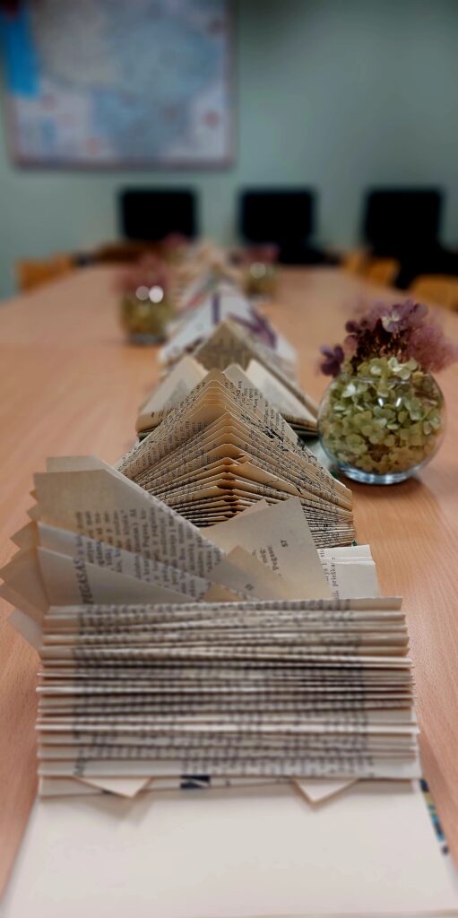 Nuotraukoje matome stalą, papuoštą įdomiu ir kūrybingu dekoru iš sulankstytų senų knygų puslapių, kurie suformuoti į piramidės formas. Kiekvienas puslapis yra tvarkingai sulankstytas ir išdėliotas, sukurdamas išskirtinį vizualinį efektą. Stalo centre matome stiklainį su džiovintais augalais, kurie papildo šį minimalistinį ir elegantišką dekorą. Iš užstalės matosi žemėlapis ir kiti stalai su kėdėmis, kas rodo, kad ši erdvė galbūt yra naudojama susitikimams ar kūrybinėms veikloms.