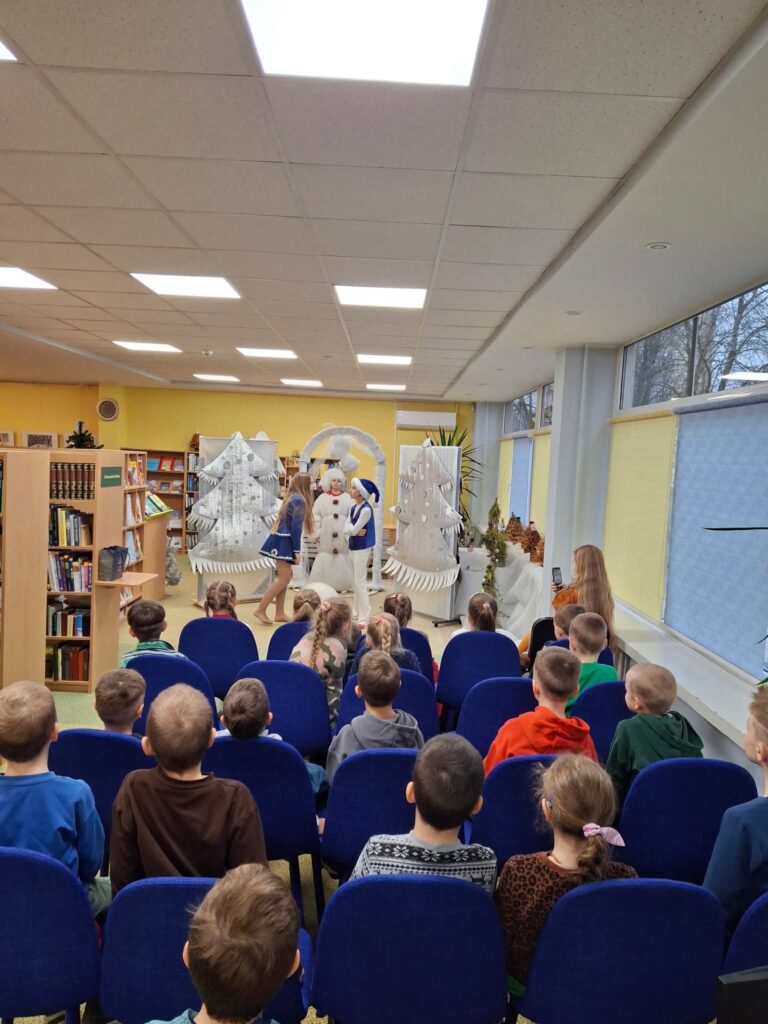 Bibliotekoje vyksta vaikams skirtas šventinis spektaklis: priešais sceną sėdi vaikai mėlynose kėdėse, o scenoje – personažai su žiemos kostiumais ir dekoracijos su eglėmis.