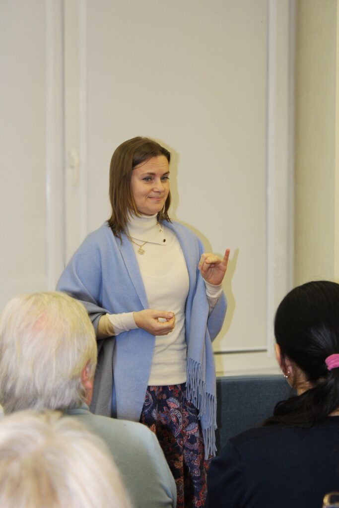 Moteris veda užsiėmimą ar paskaitą patalpoje, stovėdama priešais sėdinčius dalyvius ir gestikuliuodama ranka.