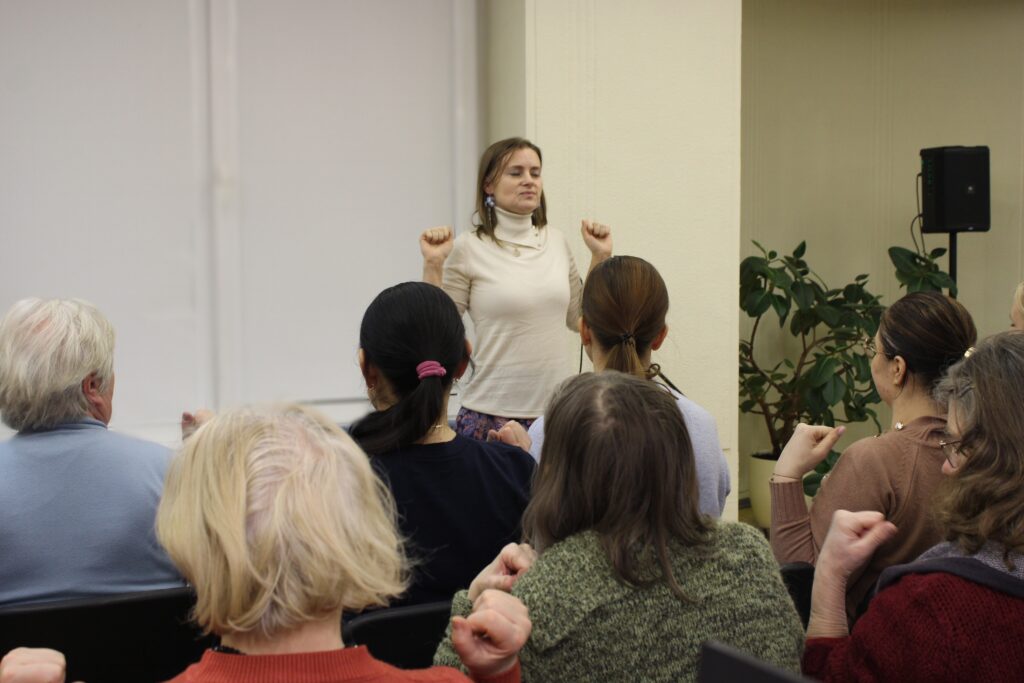 Moteris veda užsiėmimą auditorijai – stovi priešais klausytojus pakeltomis, sulenktomis rankomis, tarsi demonstruodama pratimą ar kvėpavimo techniką. Dalyviai sėdi eilėmis ir atkartoja jos judesius. Aplinka primena paskaitą ar saviugdos dirbtuves, matyti augalas ir garso kolonėlė.