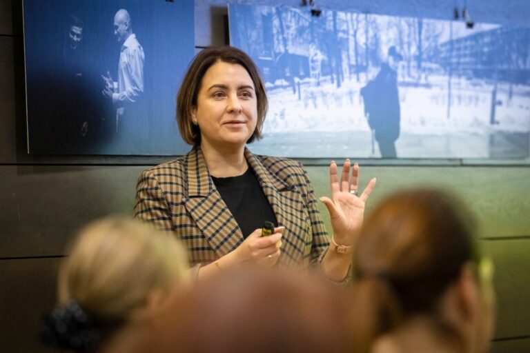 Moteris kalba išskėtusi rankos pirštus ant sienos kabančių nuotraukų fone priešais renginio auditoriją