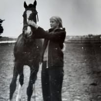 Juodai balta nuotrauka: lauke stovinti moteris švelniai laiko arklį už kamanų, aplink atvira gamta.