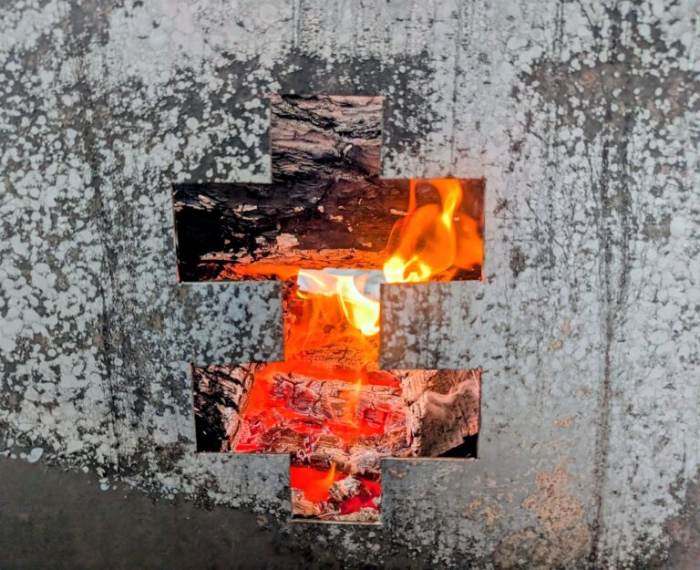 Per kryžiaus formos metalinę angą matyti degančios malkos ir žarijos, liepsnos švyti oranžine spalva pilkame, suodžiais padengtame paviršiuje.“