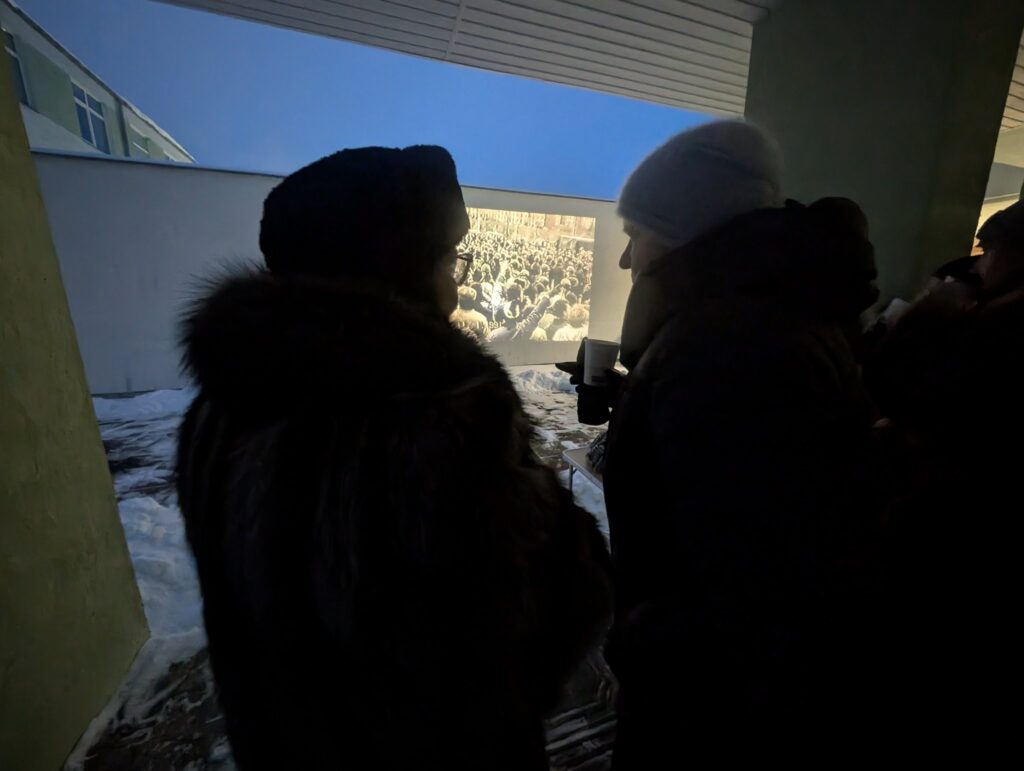 Žiemos vakarą lauke stovintys žmonės su kepurėmis ir paltais stebi ant pastato sienos projektuojamą archyvinį vaizdo įrašą su minia žmonių.